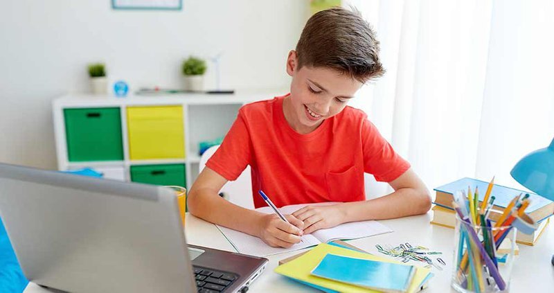 boy with red shirt sitting at desk in front of a laptop, notebooks and books
