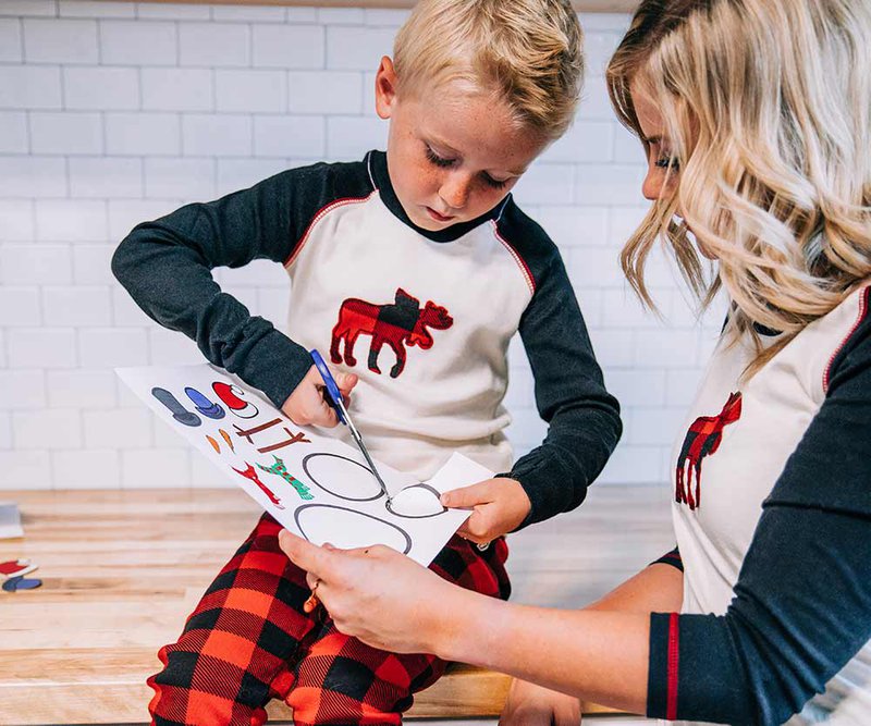 child using scissors to cut out a paper snowman with an adult supervising while wearing matching moose pajamas