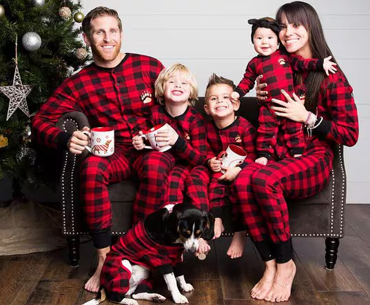 matching buffalo plaid onesies worn by a family of five and a dog, all seated posing on a small loveseat next to a Christmas tree