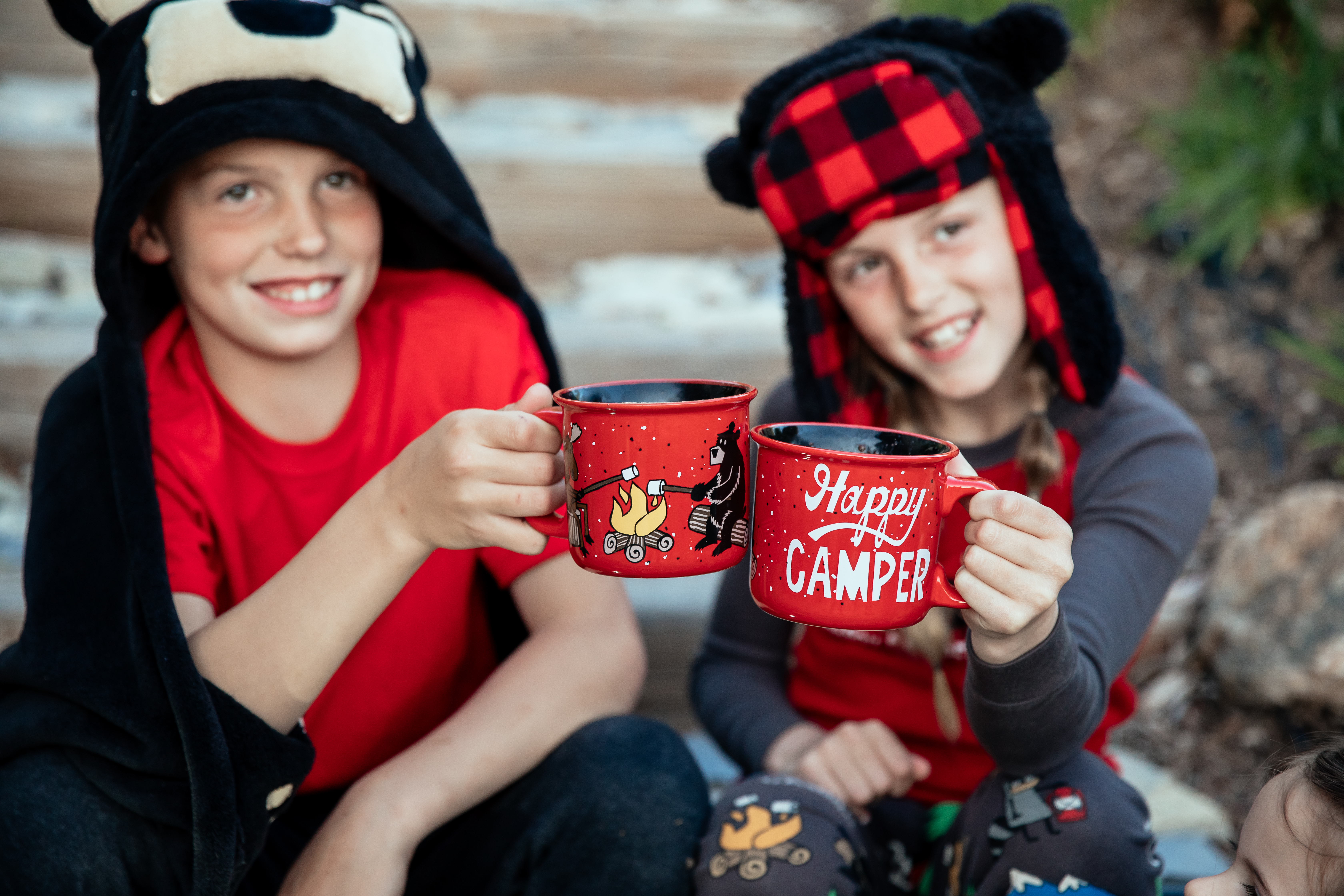 two kids shown toasting camping mugs wearing a hooded blanket and buffalo plaid cap outdoors