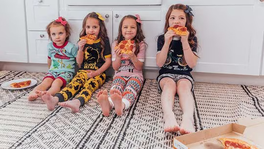 four kids wearing LazyOne pajamas eating pizza while sitting together on the floor