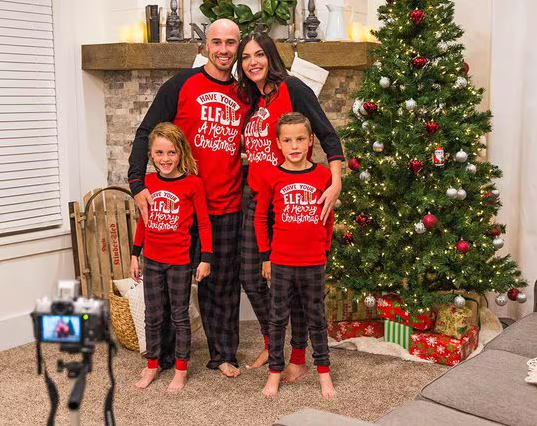 matching Christmas pajamas worn by a family of four taking a photo with a digital camera while standing next to a Christmas tree